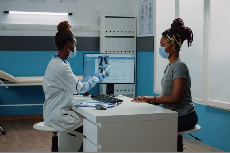 Medic holding radiography for examination with patient during coronavirus pandemic. Doctor explaining x ray scan results and diagnosis to woman, wearing face masks for protection.